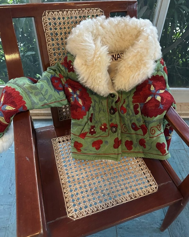 The Afghan Coat Coco No. 2 by Lunaré – lifestyle detail shot, green coat with red hand-embroidered floral pattern and cream shearling collar, draped on a rattan chair.