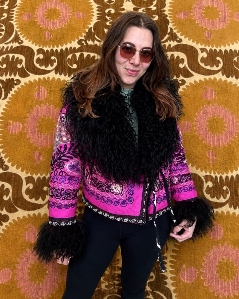 The Afghan Coat Martina No. 2 by Lunaré – lifestyle shot on model, hand-embroidered stylized animal motif in orange, cream and grey on a bright pink base, black shaggy shearling collar and cuffs.
