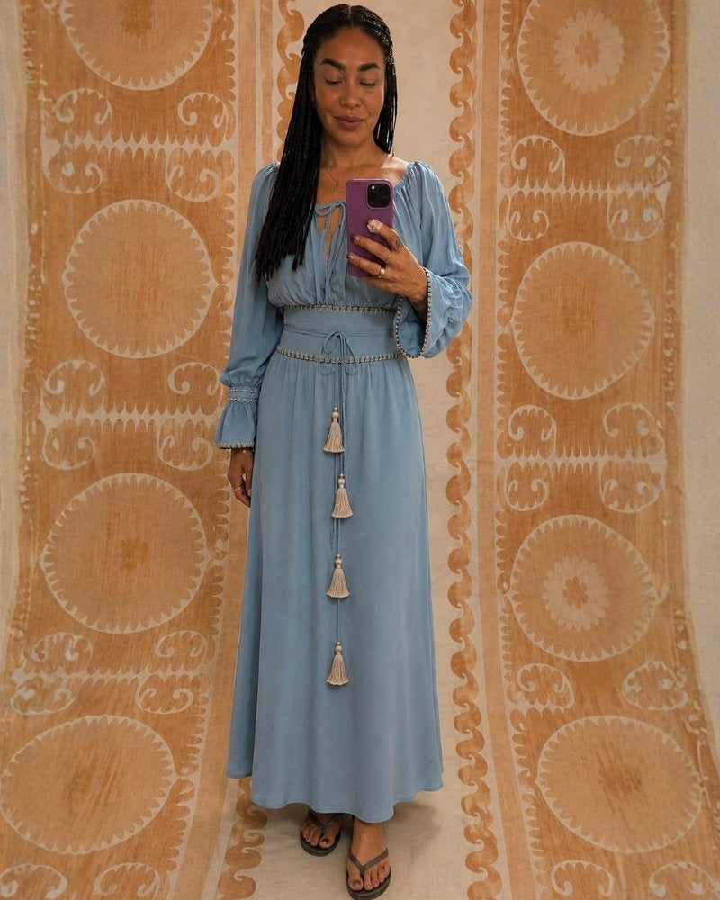 Sanaya Kim Dress by Lunaré – long-sleeve style on model, dusty blue maxi dress with smocked waist and tasseled drawstring detail.