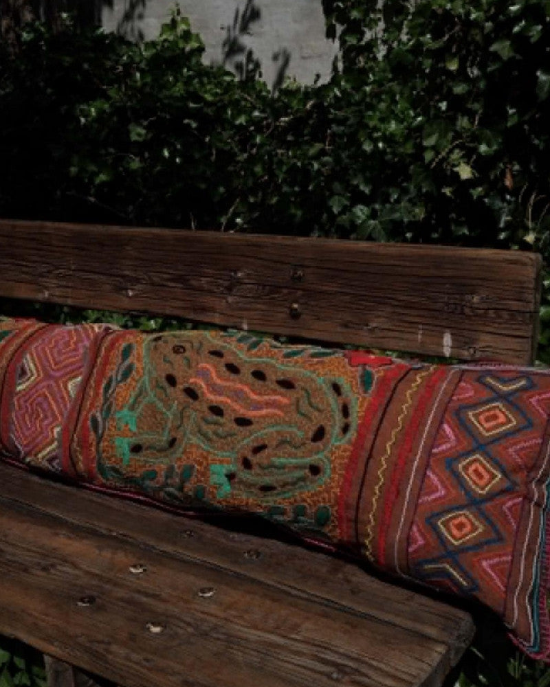 Hand-Embroidered Shipibo Cushion No. 17 by Lunaré – lifestyle shot outdoors on a wooden bench, long bolster shape with red and orange geometric embroidery.