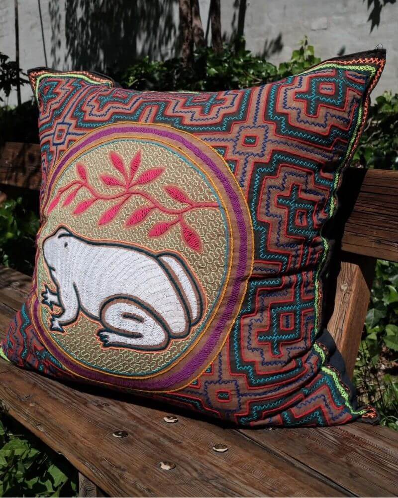 Hand-Embroidered Shipibo Cushion No. 3 by Lunaré – lifestyle shot outdoors on a wooden bench, square shape with terracotta geometric embroidery and central animal motif.