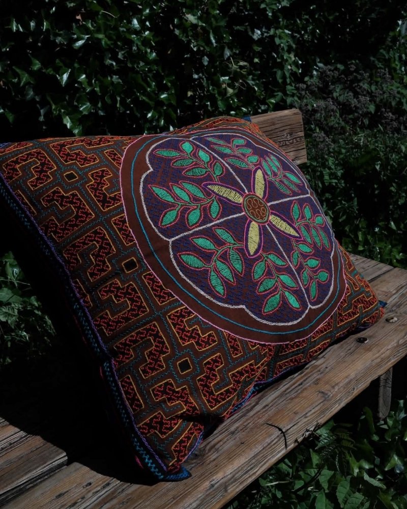 Hand-Embroidered Shipibo Cushion No. 2 by Lunaré – lifestyle shot outdoors on a wooden bench, square shape with plum geometric embroidery and central green leaf medallion.