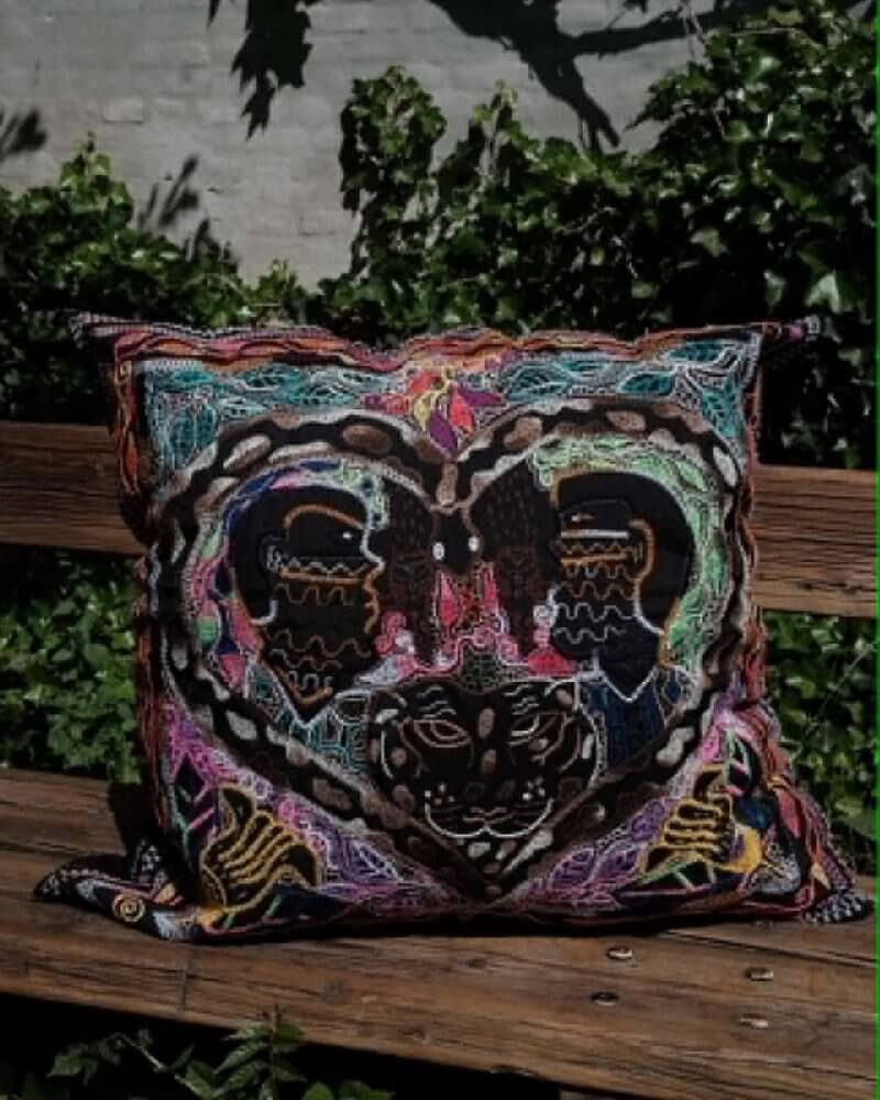Hand-Embroidered Shipibo Cushion No. 1 by Lunaré – lifestyle shot outdoors on a wooden bench, square shape with dark multicolor embroidered symmetrical face motif.