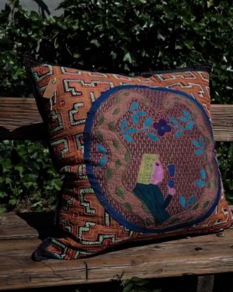 Hand-Embroidered Shipibo Cushion No. 6 by Lunaré – lifestyle shot outdoors on a wooden bench, square shape with rust geometric embroidery and central figure motif.