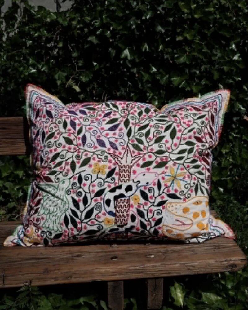 Hand-Embroidered Shipibo Cushion No. 5 by Lunaré – lifestyle shot outdoors on a wooden bench, rectangular shape with cream and pink floral bird embroidery.