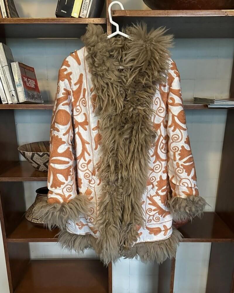 The Afghan Coat Jasmia No. 1 by Lunaré – lifestyle shot, cream coat with rust hand-embroidered floral swirl pattern and taupe shaggy shearling trim, hanging against a wooden bookshelf.
