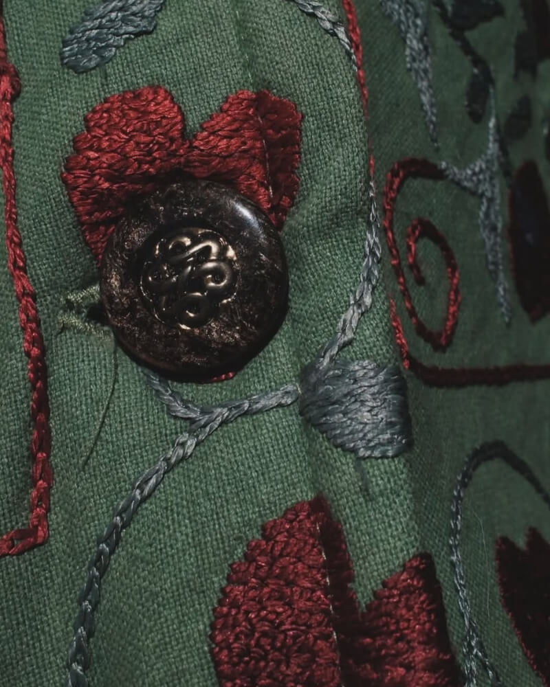 The Afghan Coat Coco No. 2 by Lunaré – close-up detail of a decorative front button and the burgundy hand-embroidered floral pattern on the sage-green textile.