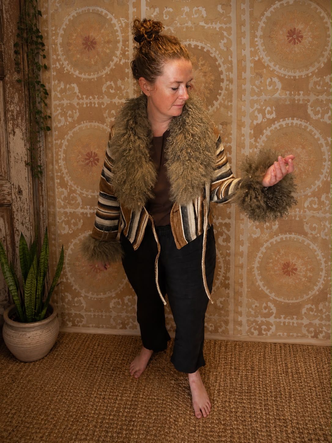 Karen Coat, worn by a model, in a striped brown and gold pattern with a shaggy brown shearling collar and cuffs, paired with black trousers.