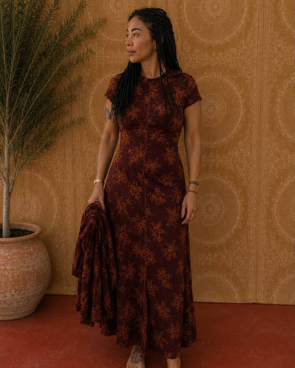 Woman wearing the Luna Dress in burgundy floral print, holding a matching jacket, standing against a patterned wall.
