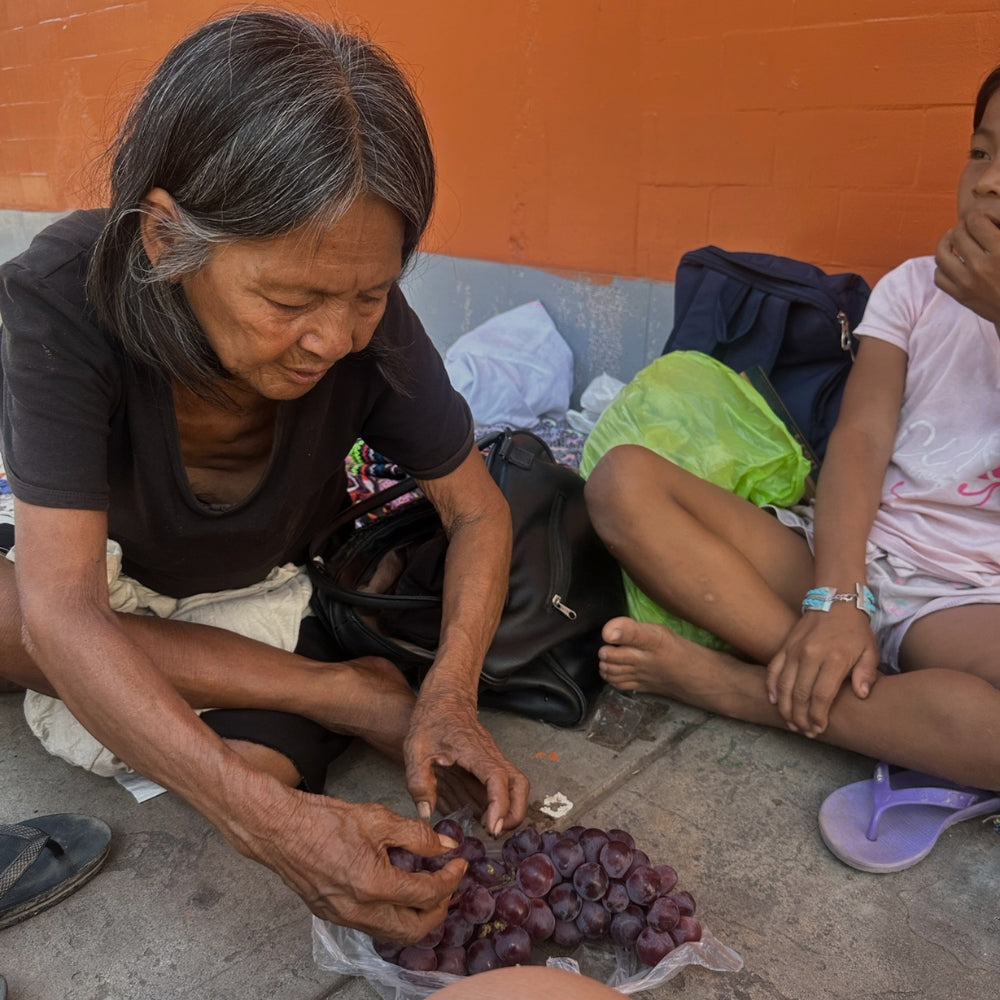 An older woman sitting on the ground sorting grapes with a child seated beside her, part of Lunaré's behind-the-scenes production photos.
