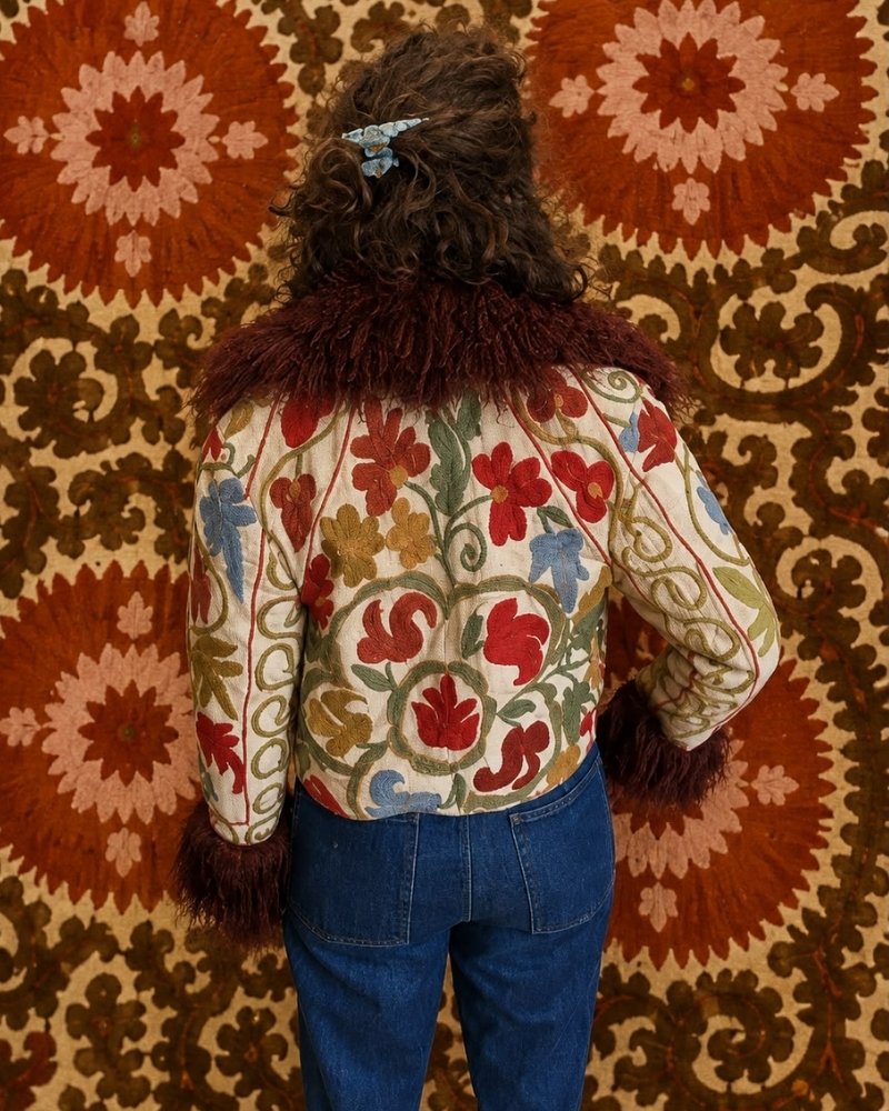 The Afghan Coat Martina No. 12 by Lunaré – back view on model, cream jacket with hand-embroidered floral vine pattern in rust, olive and blue, brown shaggy shearling collar.