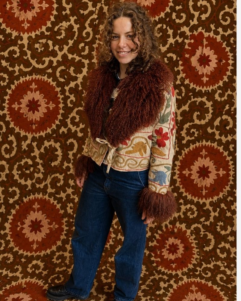 The Afghan Coat Martina No. 12 by Lunaré – on model, cream jacket with rust hand-embroidered floral pattern and brown shaggy shearling collar and cuffs, worn open over jeans.
