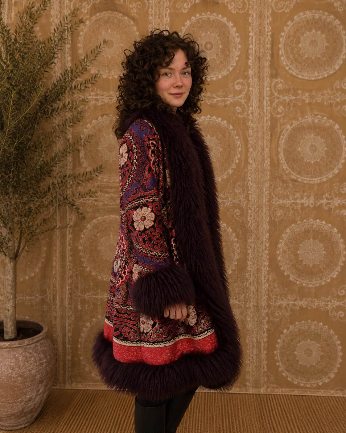 The Afghan Coat Ragna No. 4 by Lunaré – lifestyle photo of a woman wearing the mid-calf coat with red, black, cream and violet hand-embroidered pattern and burgundy shearling trim, standing indoors.