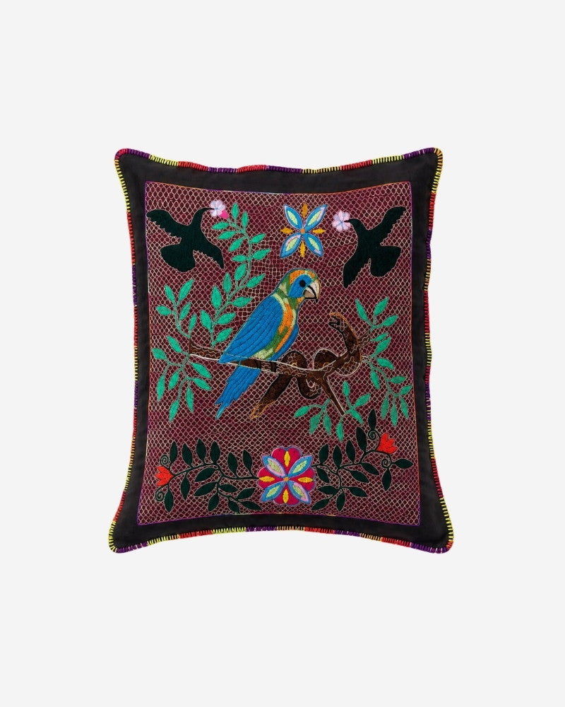 Hand-Embroidered Shipibo Cushion No. 9 in maroon, with a central turquoise parrot on a branch, two black bird silhouettes above and colourful flower medallions among green leaf foliage.