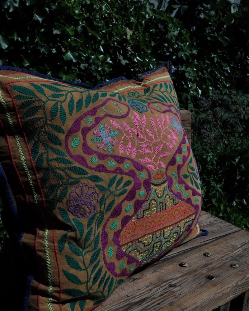 Hand-Embroidered Shipibo Cushion No. 13 by Lunaré – lifestyle shot outdoors on a wooden bench, square shape with tan and gold floral vine embroidery in pink and green.