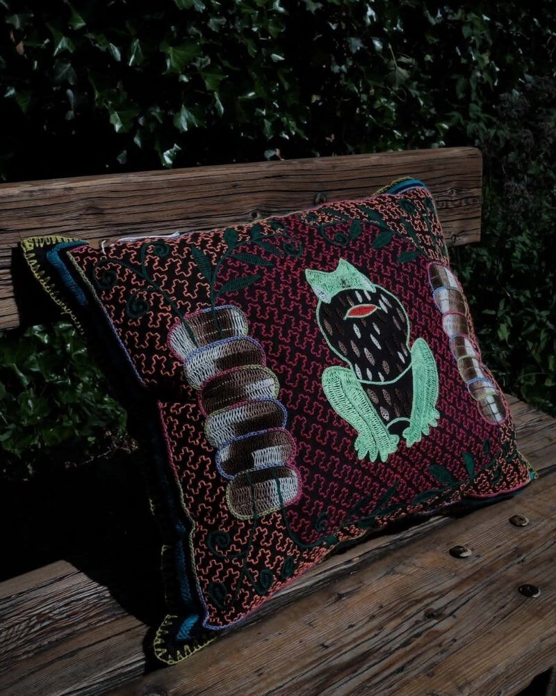 Hand-Embroidered Shipibo Cushion No. 14 by Lunaré – angled lifestyle shot outdoors on a wooden bench, maroon background with green and brown figure motif embroidery.