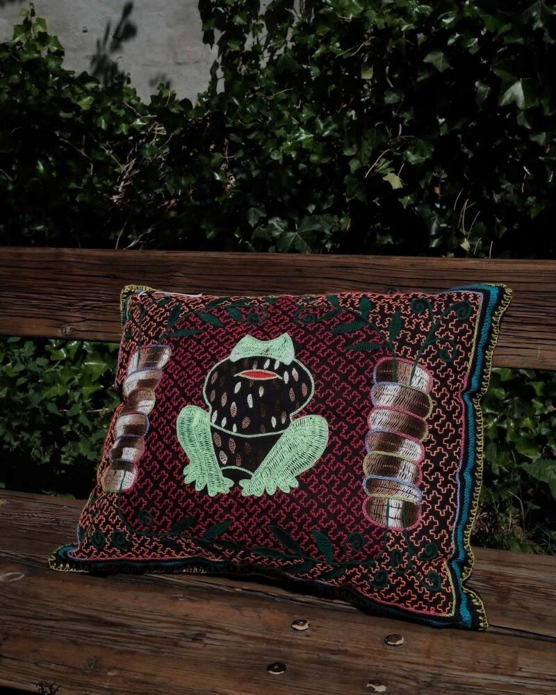 Hand-Embroidered Shipibo Cushion No. 14 by Lunaré – lifestyle shot outdoors on a wooden bench, maroon background with green and brown figure motif embroidery.