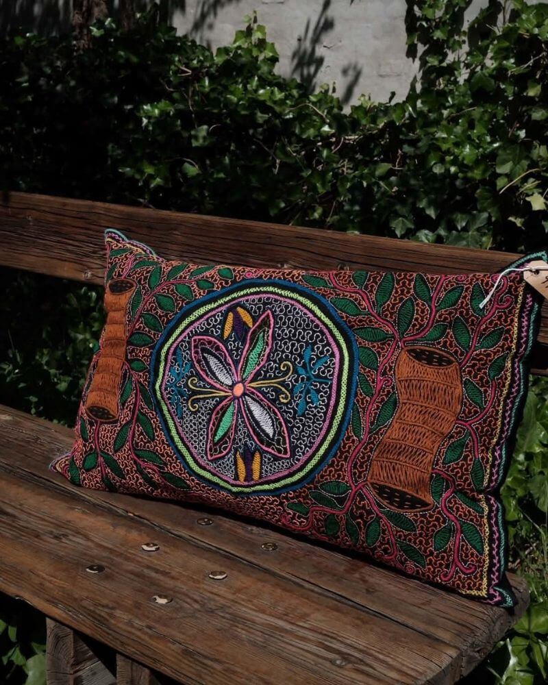 Hand-Embroidered Shipibo Cushion No. 18 by Lunaré – lifestyle shot outdoors on a wooden bench, rectangular shape with terracotta border and colorful floral medallion embroidery.
