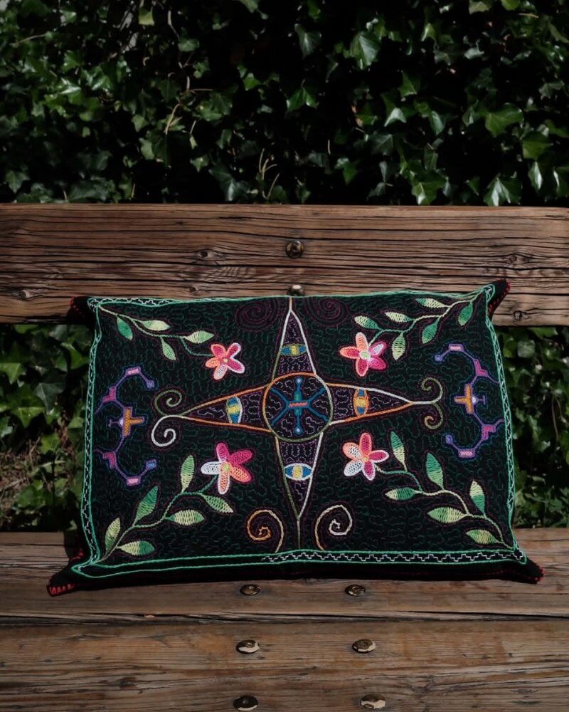 Hand-Embroidered Shipibo Cushion No. 19 by Lunaré – lifestyle shot outdoors on a wooden bench, rectangular shape with black background and colorful floral star embroidery.