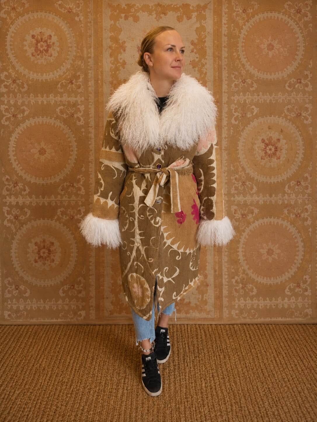 Silje Coat, worn by a model, in tan with an ornate embroidered scroll pattern and pink floral accents, a cream shearling collar and cuffs, and a tie waist closure.