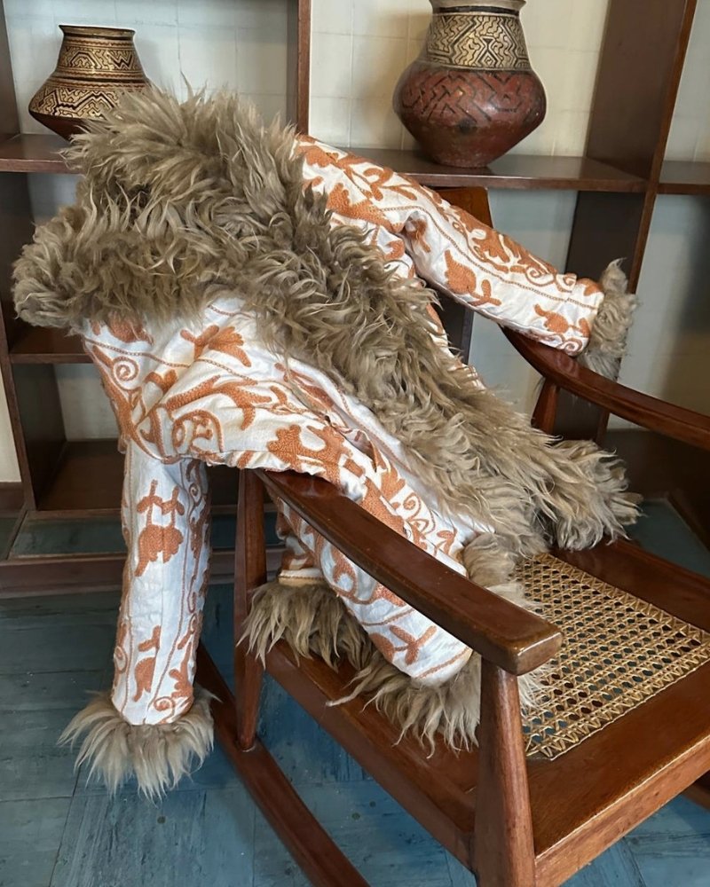 The Afghan Coat Jasmia No. 1 by Lunaré – lifestyle shot, cream coat with rust hand-embroidered floral swirl pattern and taupe shaggy shearling trim, draped over a wooden rocking chair.