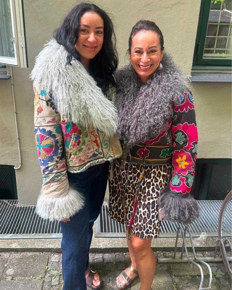 The Afghan Coat Martina No. 5 by Lunaré – lifestyle shot, two models wearing different Martina coats: cream with rainbow floral embroidery and grey shaggy shearling trim, and black with pink floral embroidery and grey shearling collar.